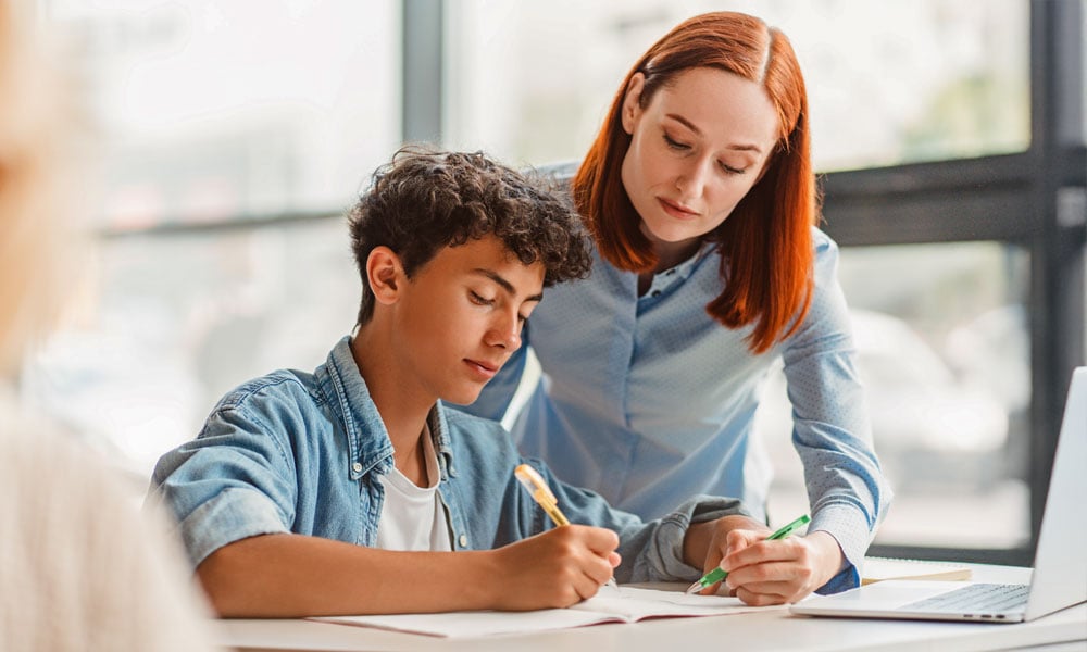 FUNDACAO-ROGE-matriz-FUNDAÇÃO ROGE: professora auxiliando um aluno que está sentado à mesa, escrevendo em seu caderno de uma das matérias da matriz curricular de sua escola-ensino-medio-e-tecnico