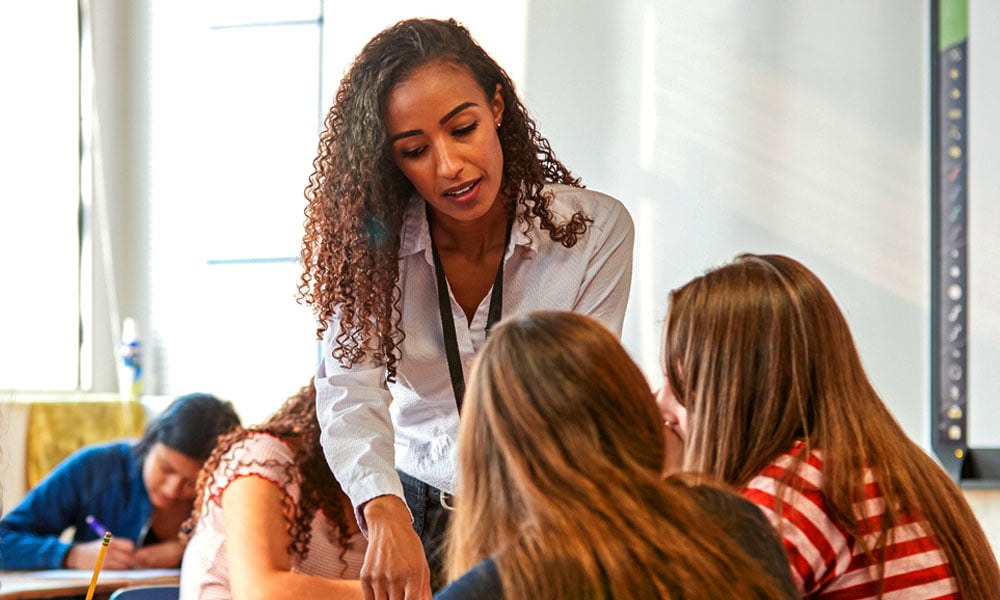 FUNDAÇÃO ROGE: professora apoiada na mesa onde estão sentados os alunos, conversando com eles, para fornecer orientação profissional de forma próxima e personalizada.